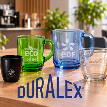 Cinq verres, tasse et mug Duralex noirs, transparents et colorés, avec des logos, disposés sur une table en bois.